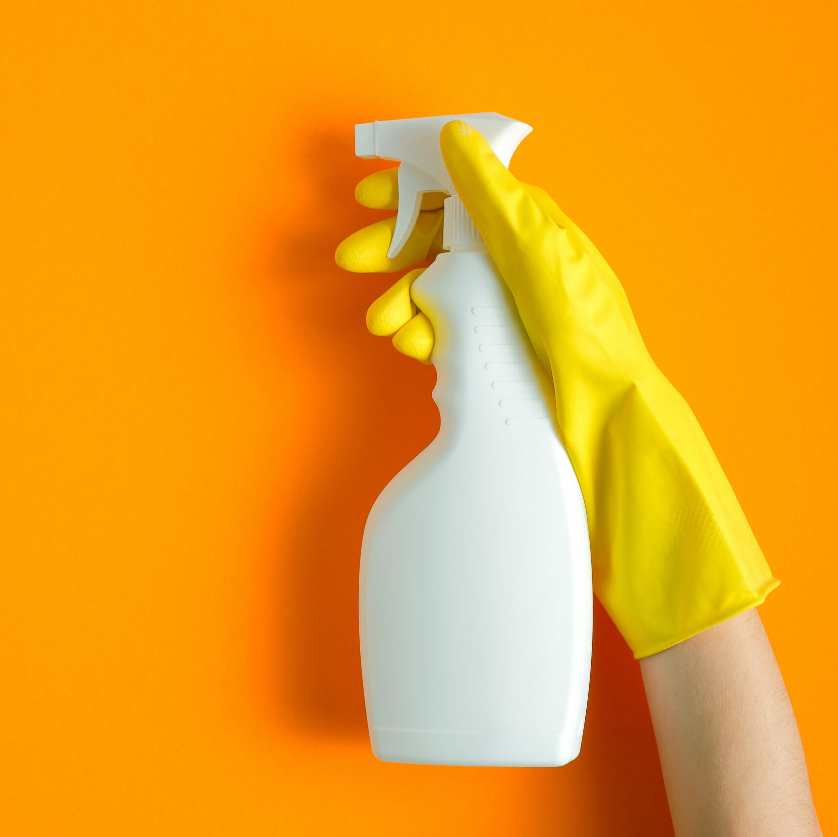 Hand in yellow rubber glove holding plastic spray bottle with cleaning detergent on an orange background