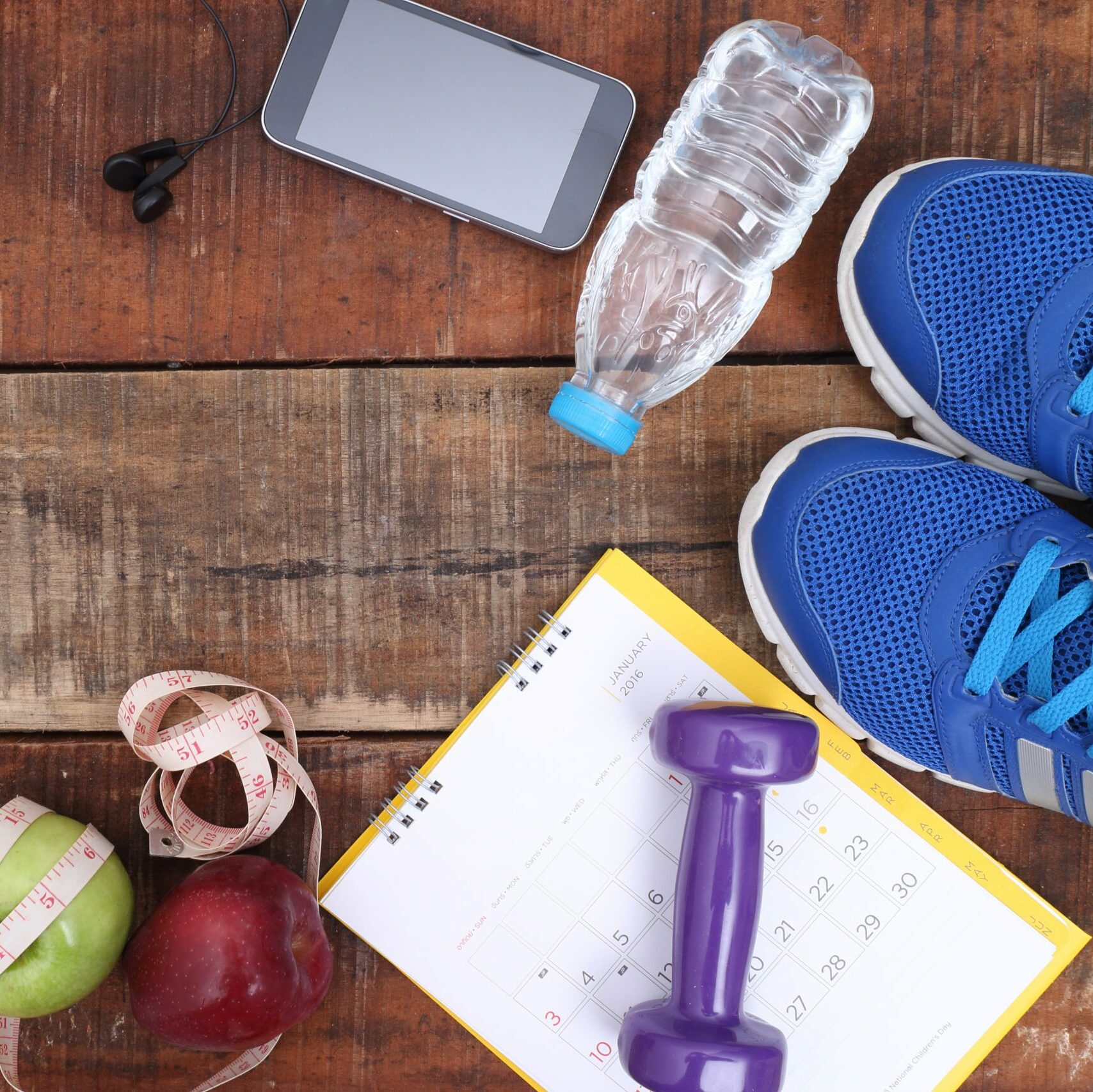 Tennis shoes, bottled water, a cell phone, a hand weight, apples, measuring tape, and a workout calendar on a wooden floor