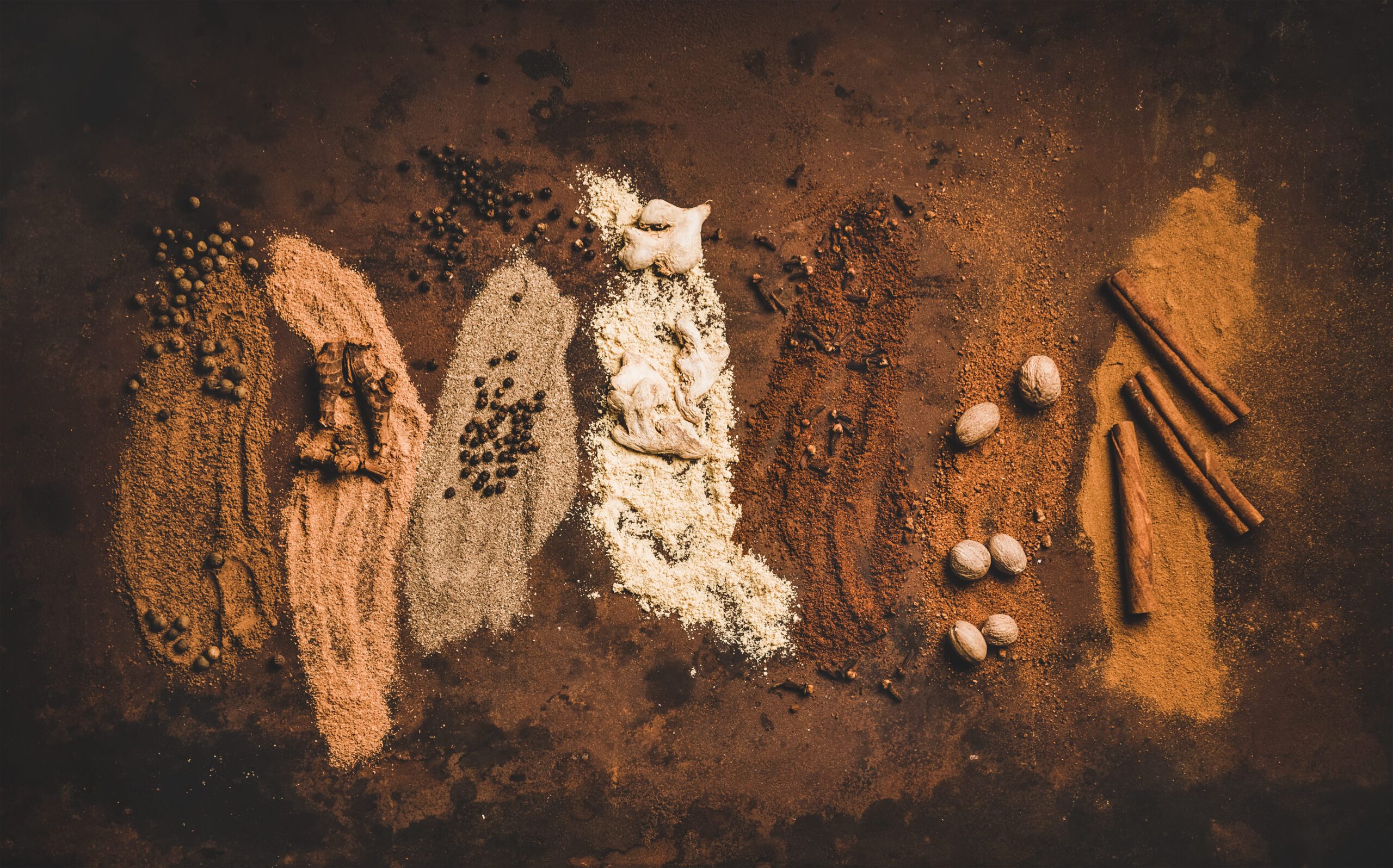 Flat-lay of black pepper, ginger, cloves, nutmeg, cinnamon, allspice, and alpinia root over a dark rusty table background