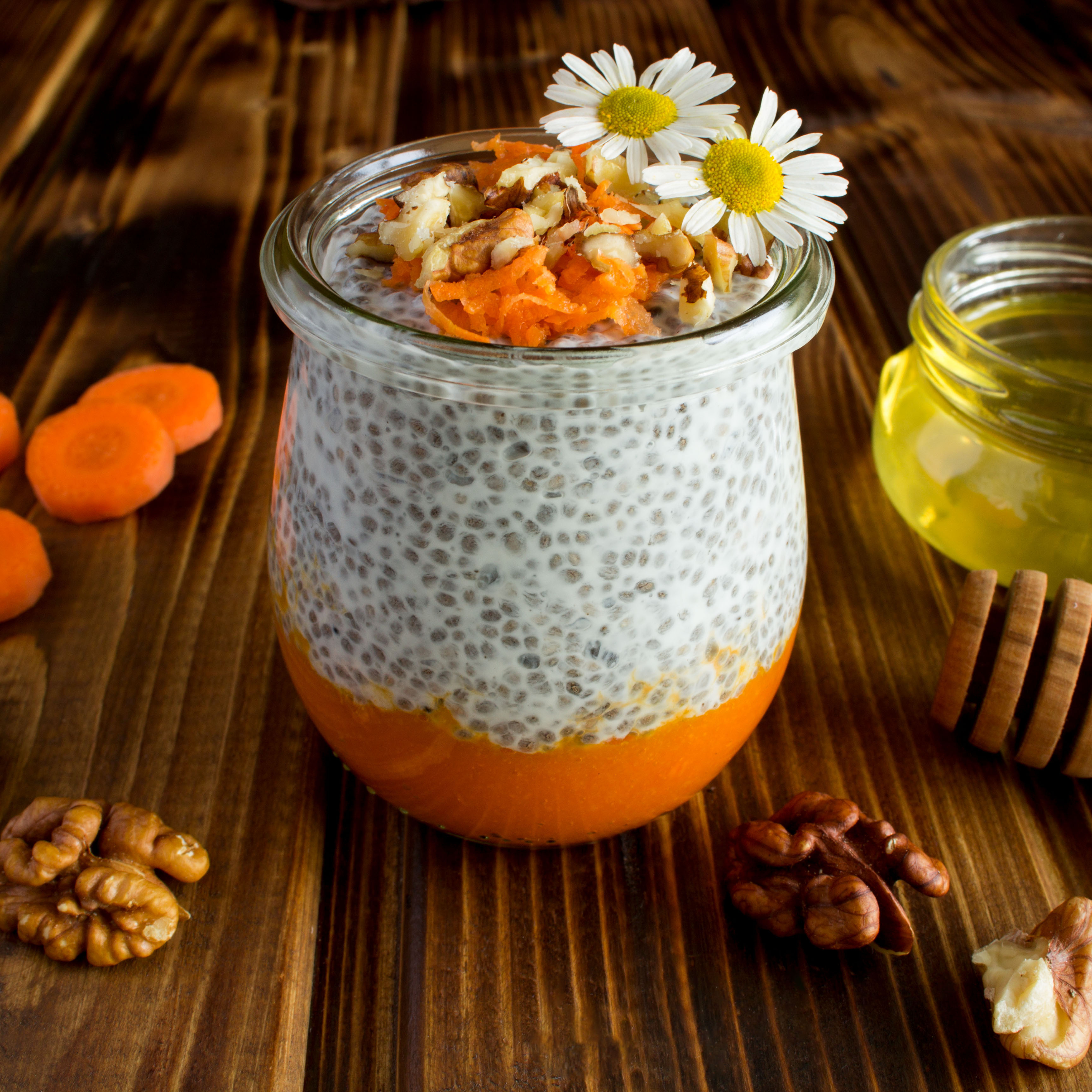 Small glass jar with an orange layer of carrot at the bottom followed by a layer of chia seed pudding. The dish is topped with nuts and two small flowers.