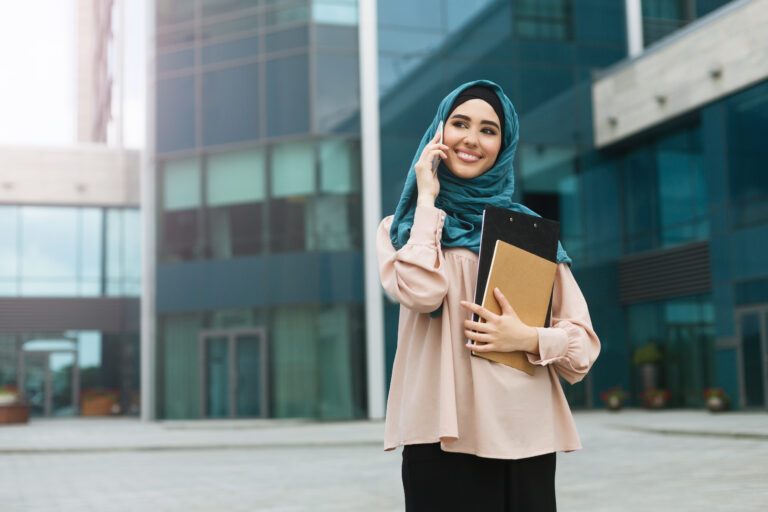 Beautiful arabic businesswoman wearing hijab talking by phone and holding folders near office building. Equal opportunities concept