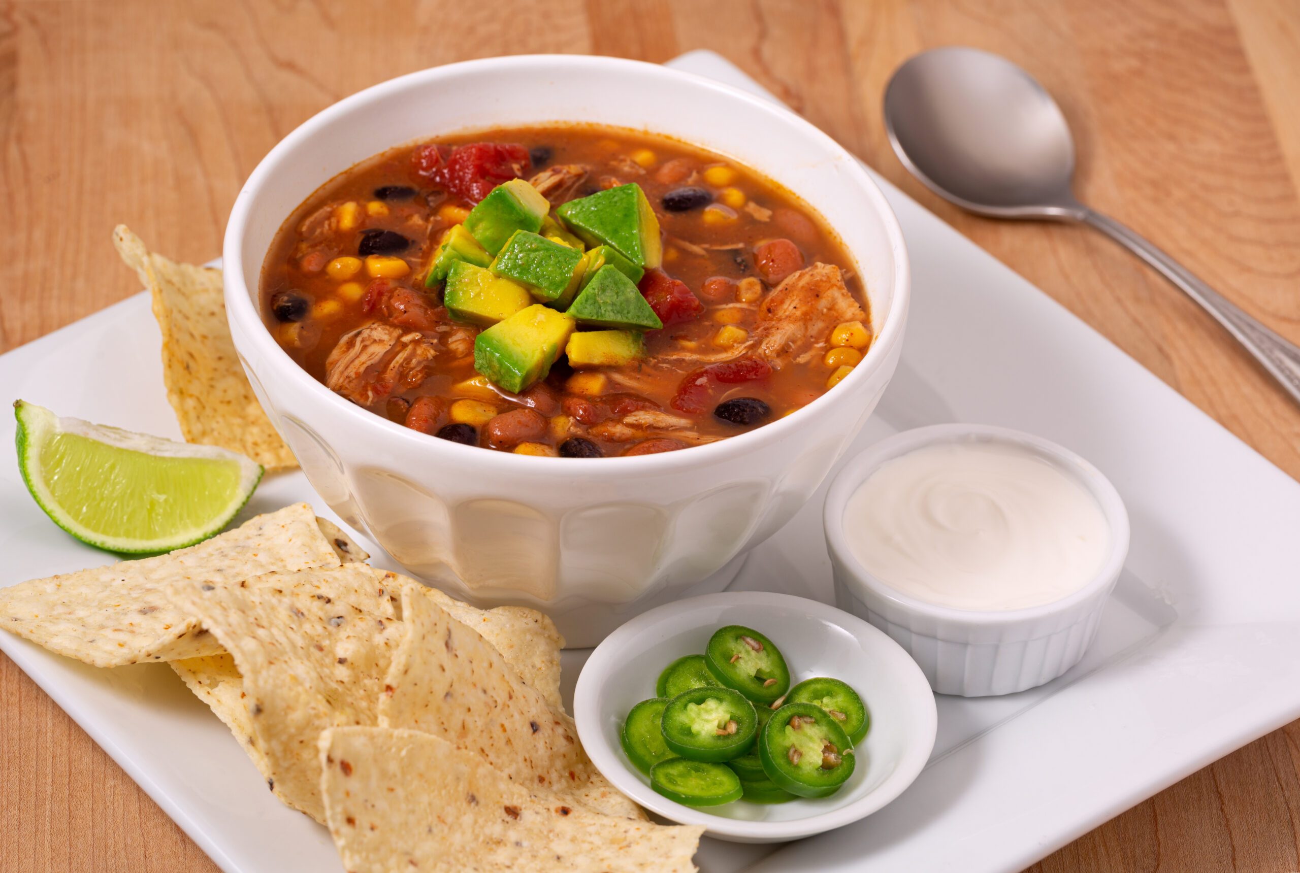 Delicious bowl of chicken taco soup garnished with avocado with sides of chili peppers, sour cream and tortilla chips