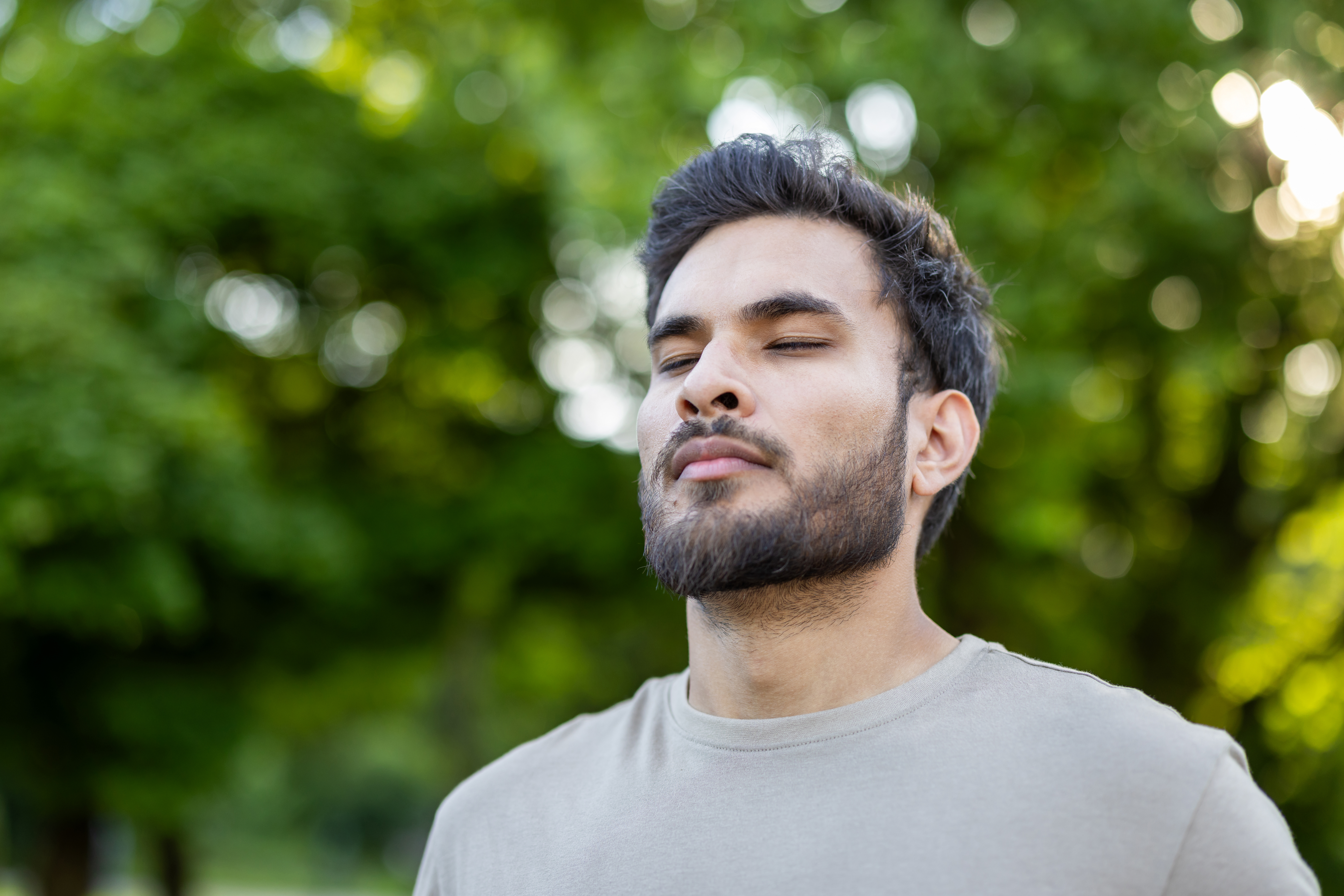 Young man stands with closed eyes embracing tranquility in serene outdoor setting. Surrounded by lush green trees, he reflects calmness and peace in natural environment.